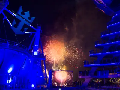 Die Harmony of the Seas bei Nacht mit spektakulärem Feuerwerk am Himmel. Das Schiff ist in leuchtendes Blau getaucht, sein Royal Caribbean Logo ist gut erkennbar. Eine große Leinwand zeigt ebenfalls Feuerwerksbilder, während Passagiere das Spektakel beobachten.