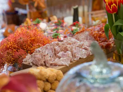 Ein appetitlich angerichtetes Buffet ist zu sehen, mit Platten voller Wurst- und Salamiaufschnitt, kleinen Vorspeisen in Gläsern und einem großen roten Blumenarrangement. Die Szene ist hell beleuchtet und detailreich dargestellt.