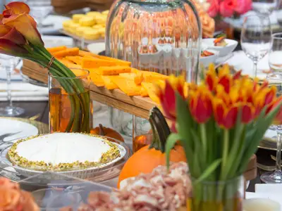 Ein festlich gedeckter Tisch mit einem Büffet aus Käseplatten, einem runden Kuchen mit Pistazienrand und herbstlicher Blumendekoration. Rote und orangefarbene Tulpen sowie Calla-Lilien sind zu sehen.