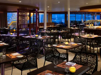 Abendansicht eines Restaurants auf der Harmony of the Seas. Viele gedeckte Tische mit dunklen Stühlen, im Hintergrund große Fenster mit blauem Licht und ein Buffetbereich.