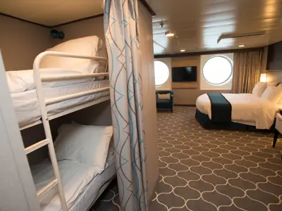 Geräumige Kabine der Harmony of the Seas mit Etagenbett, Doppelbett und runden Fenstern. Ein TV, Sessel und Schreibtisch sind im Raum. Gemusterter Teppichboden.