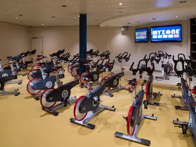 Indoor-Cycling-Studio auf der Harmony of the Seas. Viele schwarze LifeCycle-Bikes stehen auf gelbem Boden. Im Hintergrund zwei Monitore mit 'MYZONE'.