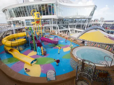 Das Splashaway Bay Wasserpark der Harmony of the Seas. Bunte Rutschen, Wasserspiele, Whirlpool und spielende Kinder sind sichtbar. Moderne gläserne Schiffaufbauten im Hintergrund.