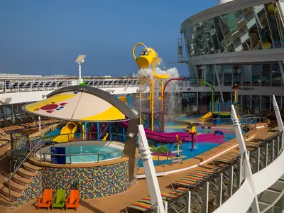 Farbenfroher Wasserpark auf dem Sonnendeck der Harmony of the Seas: Wasserrutschen, kippender Eimer, Whirlpool und Liegestühle. Rechts eine Glasfassade des Schiffs.
