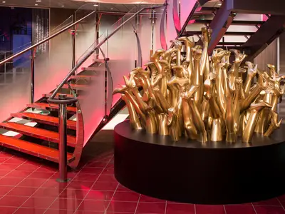 Innenbereich der Harmony of the Seas: Rotes Treppenhaus mit Glasgeländer. Eine goldene Skulptur stilisierter Beine steht auf einem schwarzen Podest.