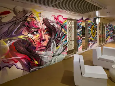 Korridor der Harmony of the Seas mit bunter Graffiti-Wandmalerei, die ein Frauengesicht und Blumen zeigt. Weiße Sessel stehen auf dem Teppichboden.