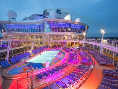 Pooldeck der Harmony of the Seas bei Dämmerung, beleuchtet mit roten, violetten und blauen Lichtern. Pool, Liegen und verglaste Aufbauten sind sichtbar.