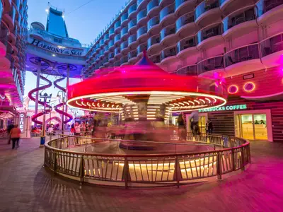 Der belebte Boardwalk auf der Harmony of the Seas bei Nacht. Ein drehendes Karussell und eine rote Rutsche sind sichtbar. Die Szene ist von pinker Beleuchtung geprägt.
