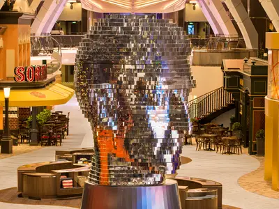 Ansicht der Royal Promenade an Bord der Harmony of the Seas. Im Zentrum steht eine große, spiegelnde Skulptur. Rechts und links sind Restaurants und Cafés mit Tischen und Stühlen zu sehen.