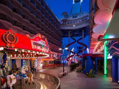 Abendliche Promenade der Harmony of the Seas. Zu sehen sind ein Karussell, beleuchtete Geschäfte, Restaurants und eine rote Wasserrutsche. Der Schiffsname 'Harmony of the Seas' ist oben sichtbar.