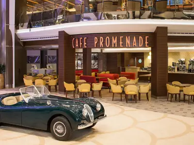 Die Royal Promenade der Harmony of the Seas mit einem dunkelgrünen Oldtimer vor dem Cafe Promenade. Gelbe Stühle und eine Kaffeebar sind sichtbar.