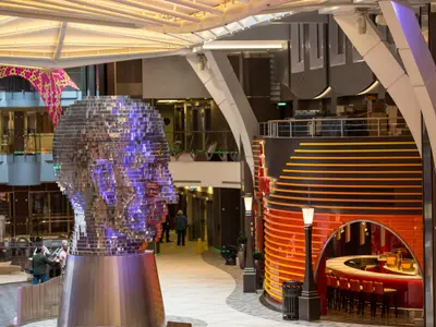 Innenansicht der Harmony of the Seas: Eine glitzernde, spiegelnde Kopfskulptur steht im Vordergrund. Im Hintergrund sind eine orange-gestreifte Bar und verschiedene Ebenen des Schiffes zu sehen, beleuchtet und modern.