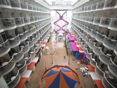Draufsicht auf das Boardwalk-Viertel der Harmony of the Seas. Viele Balkonkabinen säumen die Seiten. Eine lila Wasserrutsche und Geschäfte füllen die Promenade. Karussellzelt im Vordergrund.