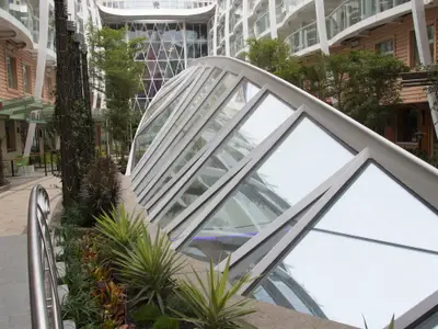 Blick in den Central Park der Harmony of the Seas: Eine begrünte Promenade mit geneigtem Glasdach und Kabinenbalkonen im Hintergrund.