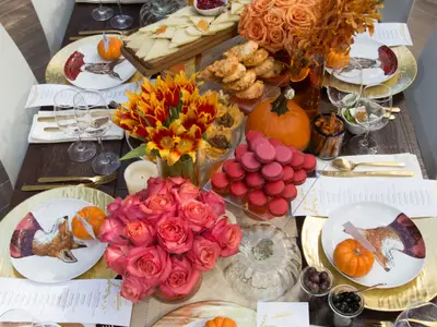 Eine festlich gedeckte Tafel für ein Friendsgiving-Essen, dekoriert mit orangefarbenen Kürbissen, Blumen, goldenem Besteck und Tellern mit Tiermotiven. Verschiedene Speisen, darunter Empanadas und rote Macarons, sind appetitlich arrangiert.