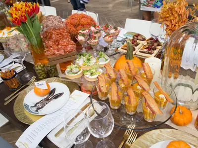 Eine lange Tafel, festlich dekoriert mit Herbstblumen und kleinen Kürbissen, bietet eine Vielfalt an Vorspeisen und Snacks. Darunter Aufschnitt, Canapés, Suppen in Gläsern mit Bacon und kleine Gebäckstücke. Gläser und Geschirr sind für Gäste bereitgestellt.