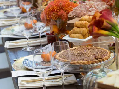 Ein herbstlich dekorierter Tisch, festlich gedeckt. Zu sehen sind ein Pekannusskuchen, frittierte Snacks, Aufschnitt, Weingläser und kleine Kürbisse.
