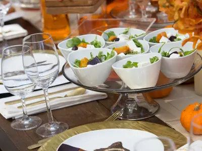 Detailaufnahme eines elegant gedeckten Tisches mit kleinen Salatschalen auf einer Etagere und leeren Weingläsern. Dekorative Kürbisse und Getränke im Hintergrund. Zeigt eine festliche Atmosphäre.