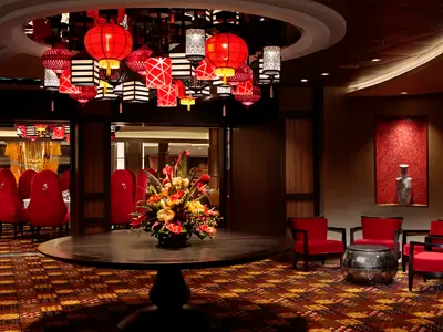Innenansicht eines asiatisch inspirierten Restaurants mit roten Samtstühlen, dunklen Holzelementen und leuchtenden Lampions an der Decke. Ein großer Blumentisch und eine Lounge-Ecke sind im Vordergrund zu sehen.