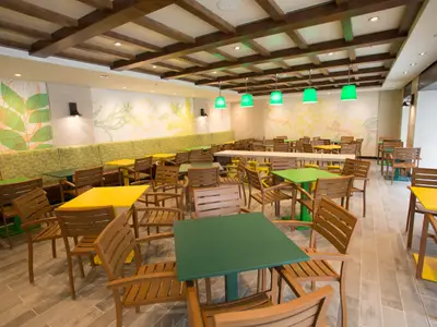 Ein großes, helles Restaurant mit zahlreichen Holzstühlen und Tischen mit grünen und gelben Platten. Die Wände sind mit floralen Mustern in Pastellfarben verziert. Grüne Hängelampen spenden Licht.