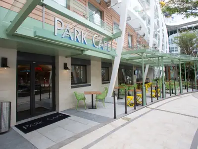 Der Eingangsbereich des Park Cafe auf der Harmony of the Seas. Es zeigt Außensitzplätze mit grünen Stühlen, Holztischen und gelben Hockern unter einer hellgrünen Glasüberdachung.