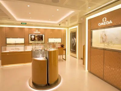 Der Omega-Shop auf der Harmony of the Seas zeigt luxuriöse Uhren und Schmuck in Vitrinen. Das elegante Geschäft ist mit Omega-Logos und warmer Beleuchtung ausgestattet. Ein grünes Ausgangsschild ist sichtbar.