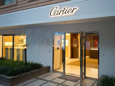 Cartier-Geschäft auf der Harmony of the Seas mit geöffneter Glastür. Beleuchtetes Logo, innen Luxuswaren in Vitrinen. Links ein Omega-Geschäft. Vorbereich mit Pflanzen und Laternen.