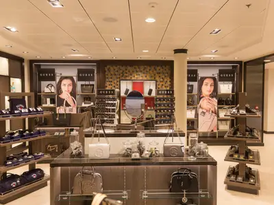 Ein stilvolles Einzelhandelsgeschäft an Bord der Harmony of the Seas zeigt Taschen, Uhren und Schmuck der Marken Guess und Hilfiger auf Holzregalen und in Vitrinen.