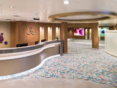 Empfangsbereich des Vitality at Sea SPA auf der Harmony of the Seas. Ein geschwungener Tresen, gemusterter Teppich und Wandbilder kennzeichnen den hellen Raum.