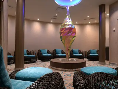 Ein luxuriöser Innenraum mit sanften Erdtönen an den Wänden und vielen blauen, plüschigen Sitzgelegenheiten in Rattansesseln. In der Mitte steht eine hohe, farbenfrohe, prismatische Skulptur auf einem runden Podest. Mehrere Säulen strukturieren den Raum. Die Beleuchtung ist sanft, mit Deckenspots.