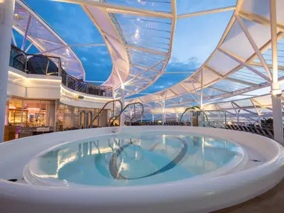 Der Whirlpoolbereich der Harmony of the Seas am Abend. Ein großer ovaler Pool unter einem geschwungenen Glasdach, umgeben von Decks, Restaurants und Liegestühlen. Der Himmel ist blau.