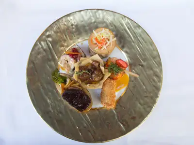 Eine goldene Servierplatte mit einem einzelnen, kunstvoll angerichteten Gericht. Es besteht aus mehreren kleinen Portionen: einem Fleischröllchen, einem Schälchen mit Schmorgericht, einem halben Brötchen mit dunkler Soße, einem frittierten Krokettenstück, Garnelen mit Mango-Soße und einem Turm aus Speisen mit Julienne-Gemüse.