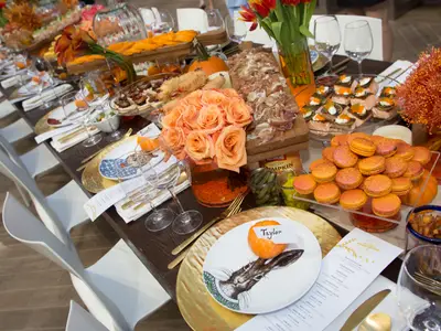 Ein langer, festlich gedeckter Tisch, beladen mit einer Vielzahl von Speisen, darunter Aufschnitt, kleine Sandwiches, Macarons und Vorspeisen. Orangefarbene Rosen, kleine Kürbisse und weitere herbstliche Dekorationen zieren die Tafel. Einzelne Platzkarten mit Namen sind sichtbar, aber es sind keine Personen anwesend.