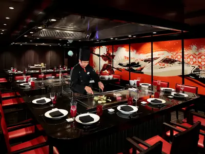 Ein Koch bereitet im Teppanyaki-Restaurant auf der Harmony of the Seas Speisen am Grill zu. Gästeplätze mit roten Stühlen und japanisch inspirierten Wandbildern prägen das Ambiente.