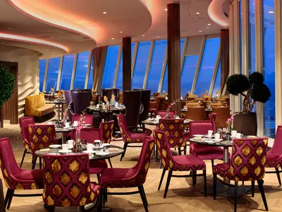 Innenansicht des eleganten Restaurants auf der harmony of the seas. Zahlreiche Tische mit rotsamtigen Stühlen sind gedeckt. Große Fensterfronten zeigen den blauen Himmel am Abend.