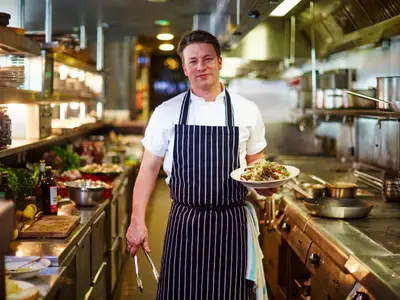Koch Jamie Oliver lächelt in einer Profiküche. Er trägt Kochjacke und Schürze, präsentiert einen Teller mit Speise. Im Hintergrund sind Küchengeräte und Regale.