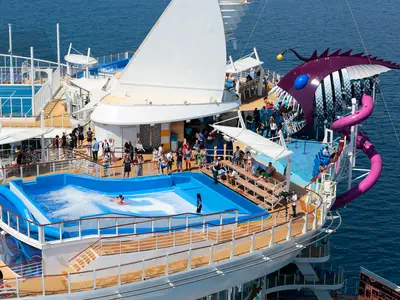 Luftaufnahme der Harmony of the Seas zeigt FlowRider, Pools, pinke Rutsche und die markante Anglerfisch-Rutsche 'The Abyss'. Zahlreiche Passagiere sind an Deck zu sehen.