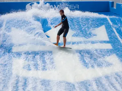 Auf der Harmony of the Seas surft eine Person in schwarzem Neoprenanzug auf dem FlowRider-Simulator. Klares Wasser spritzt auf der blauen Bahn mit weißem Royal Caribbean Logo.