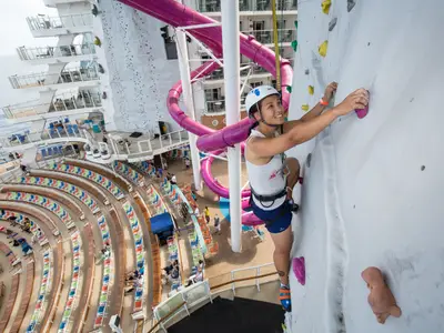 Eine lächelnde Frau klettert gesichert an der Kletterwand der Harmony of the Seas. Im Hintergrund sind lila Wasserrutschen und ein Amphitheater zu sehen.