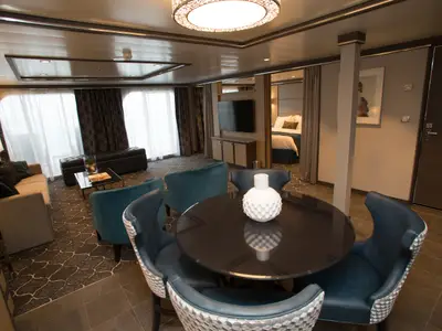 Blick in eine geräumige Suite auf der Harmony of the Seas. Man sieht einen Wohnbereich mit Sofa, Esstisch mit blauen Stühlen und das angrenzende Schlafzimmer.