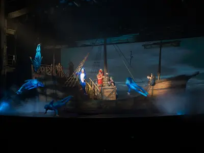 Bühnenshow der Harmony of the Seas. Akrobaten in blauen Anzügen schweben um ein großes Schiffsset. Darsteller auf dem Deck. Blaulicht und Nebel.