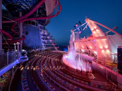 Das nächtliche AquaTheater an Bord der Harmony of the Seas zeigt eine farbenfrohe Fontänenshow. Links sind eine Kletterwand und eine rosa Wasserrutsche zu sehen, rechts das offene Meer.
