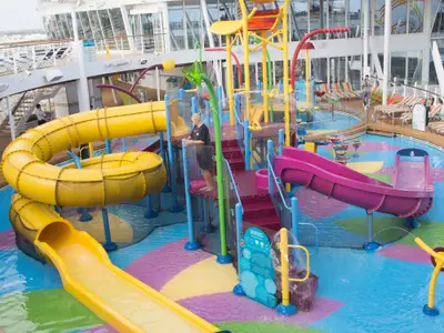 Blick auf den farbenfrohen Wasserpark "Splashaway Bay" an Deck der Harmony of the Seas. Zu sehen sind gelbe und pinke Wasserrutschen, verschiedene Sprühdüsen und Wasserbehälter in einem flachen Becken. Ein Mitarbeiter steht auf einer Plattform. Im Hintergrund sind weitere Decks und Liegestühle erkennbar.