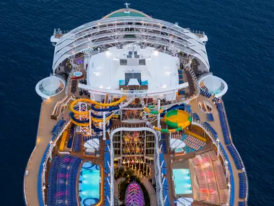 Luftaufnahme der oberen Decks der Harmony of the Seas mit bunten Wasserrutschen, mehreren Pools, vielen Liegestühlen und beleuchteten Bereichen am Abend. Das Schiff fährt auf blauem Meer.