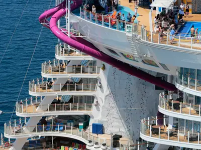 Detailansicht der Harmony of the Seas mit einer markanten magenta Wasserrutsche und einer Felswand zum Klettern. Mehrere Balkone und obere Decks mit Passagieren sind zu sehen.