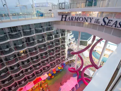 Vogelperspektive auf das offene Oberdeck der Harmony of the Seas. Mehrere Stockwerke von Balkonkabinen umrahmen eine zentrale Promenade mit bunten Sitzgelegenheiten, pinken Wasserrutschen, einem Poolbereich und Blick auf das Meer.