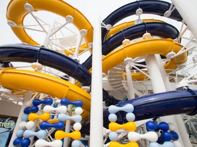 Ansicht der gelben und dunkelblauen Wasserrutschen auf dem Oberdeck der Harmony of the Seas. Weiße Stützen und eine Wendeltreppe sind zu sehen. Der Himmel ist hellgrau.
