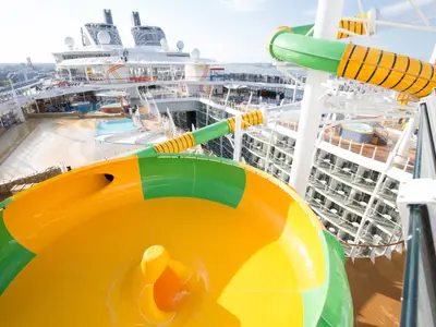 Eine gelb-grüne Wasserrutsche auf dem Oberdeck der Harmony of the Seas ist zu sehen, mit Pools, Balkonkabinen und den Schornsteinen des Schiffs im Hintergrund.