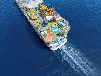 Luftaufnahme des Kreuzfahrtschiffs Hero of the Seas auf blauem Meer. Man sieht einen Wasserpark, Pools, Sportplätze und Liegeflächen auf den oberen Decks. Eine deutliche Heckwelle ist sichtbar.