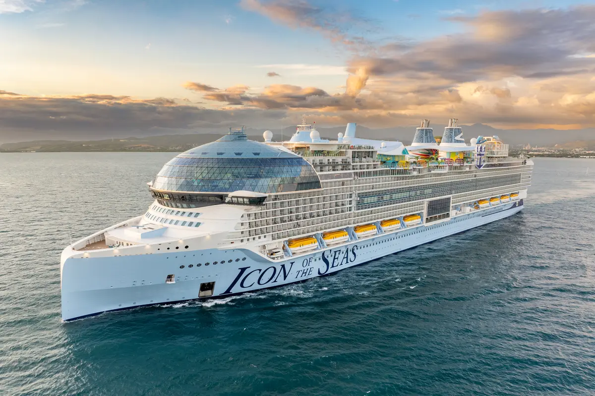 Der riesige Kreuzer Icon of the Seas von Royal Caribbean gleitet bei Sonnenuntergang auf dem Meer. Berge und eine Küstenlinie sind im Hintergrund zu sehen. Das Schiff hat eine große Glaskuppel am Bug und bunte Decksbereiche am Heck.